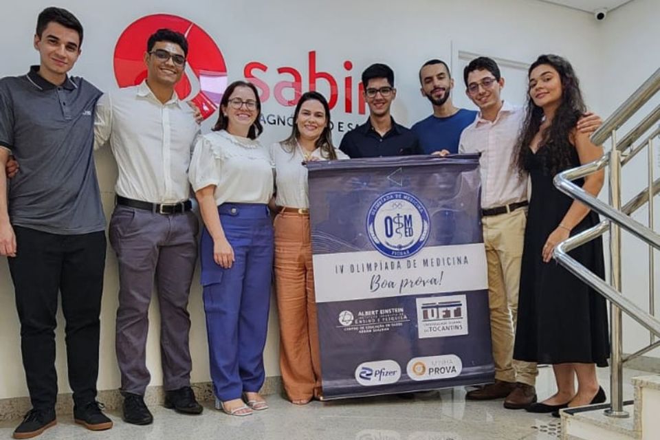 Na imagem, um grupo de alunos, professores e gestores está em pé, sorrindo para a câmera. Ao lado deles, há um banner da Olimpíada de Medicina, destacando o evento.