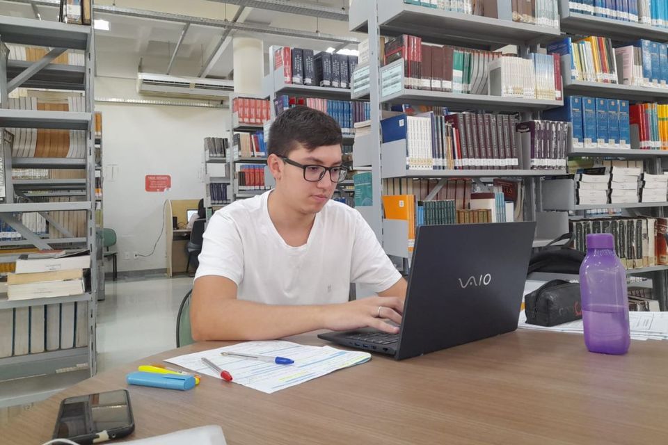 Jovem sentado em frente ao notebook, em ambiente de biblioteca, com estantes de livros ao fundo