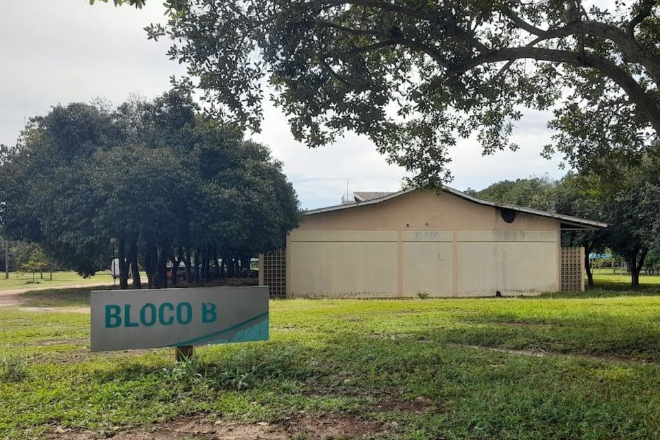A imagem mostra o Bloco B do Câmpus de Palmas da UFT, cercado por árvores e área verde. Em primeiro plano, há uma placa identificando o bloco. O prédio tem estrutura simples, fachada bege com sinais de desgaste e um telhado inclinado. O ambiente está tranquilo, sem presença de pessoas.