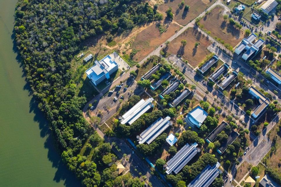 A imagem mostra uma fotografia aérea do Câmpus de Palmas da UFT. À esquerda, há uma extensa área verde margeando um grande corpo d’água. À direita, distribuem-se diversos prédios acadêmicos e administrativos, com telhados predominantemente claros, cercados por árvores e vias internas. No centro superior, destaca-se um prédio maior, isolado, com arquitetura moderna. A iluminação indica que a foto foi feita em um dia ensolarado, criando sombras alongadas sobre o terreno.
