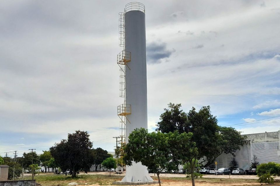 Fotografia em plano aberto mostra um reservatório metálico cilíndrico de grande porte, pintado na cor branca, instalado em posição vertical sobre uma base elevada. A estrutura possui escadas e plataformas de acesso externas em amarelo e se destaca acima das árvores ao redor. O reservatório está localizado em uma área do Câmpus de Palmas da UFT, com edificações, veículos estacionados e vegetação ao fundo. O céu está parcialmente nublado, com nuvens claras e trechos de azul visíveis.