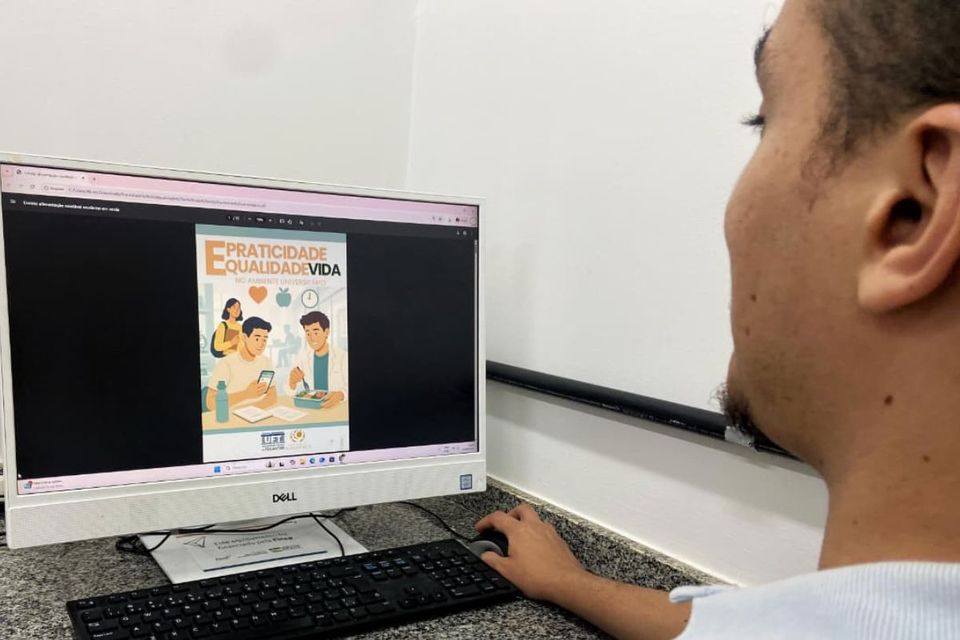 Um jovem sentado diante de um computador observa na tela a capa digital da cartilha “Praticidade e qualidade de vida no ambiente universitário”, que mostra ilustrações de estudantes se alimentando.