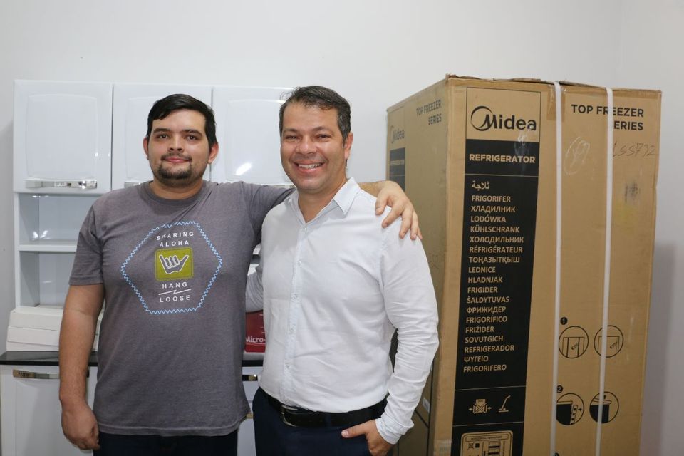 Na imagem, o estudante Thales Marques Rodrigues, de camiseta cinza, está abraçado ao diretor do Câmpus de Palmas, professor Moisés de Souza Arantes Neto, que veste camisa social branca. Ambos sorriem, posando em um ambiente interno com armários brancos ao fundo e uma grande caixa de papelão identificada com a marca “Midea”, que contém um refrigerador.