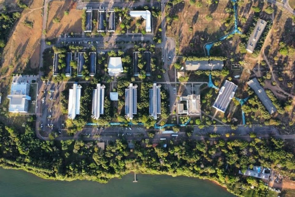 Fotografia aérea, vista de cima, do Câmpus de Palmas da Universidade Federal do Tocantins. A imagem mostra um amplo conjunto de prédios distribuídos entre áreas arborizadas, estacionamentos e vias internas. Na parte inferior da fotografia há uma extensa área de vegetação e, logo depois, um trecho do lago, com um pequeno píer avançando sobre a água. Os edifícios possuem diferentes formatos e estão interligados por caminhos e ruas. A paisagem apresenta bastante arborização, destacando a integração entre as construções, a natureza e o lago.