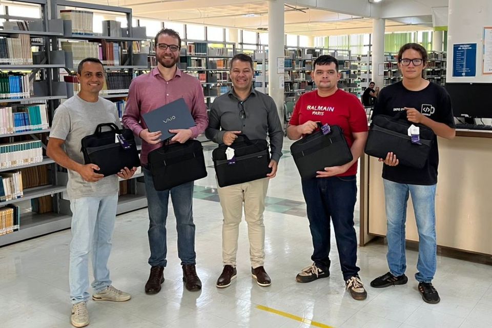Cinco homens posam sorridentes no interior da Biblioteca do Câmpus de Palmas, em meio a estantes com livros. Cada um segura uma bolsa de notebook preta, e um deles também segura um notebook aberto. Ao fundo, é possível ver mesas de estudo, janelas amplas e iluminação natural preenchendo o ambiente.