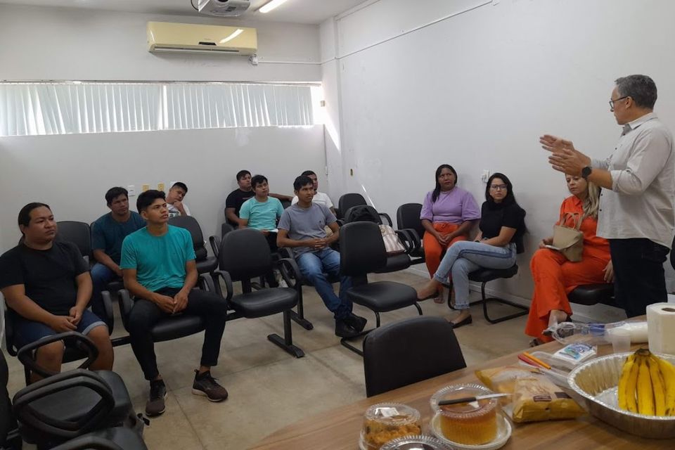 Foto de uma sala com paredes brancas e iluminação fluorescente, um grupo de cerca de 13 pessoas participa de uma reunião. A maioria está sentada em cadeiras organizadas em semicírculo, ouvindo atentamente um homem de cabelos grisalhos, que está em pé à direita da imagem, falando com gestos expressivos.