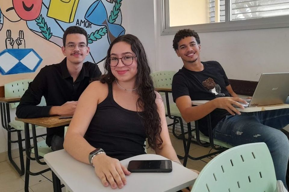 Foto com três jovens, sentados, em sala de aula, em cadeiras escolares 