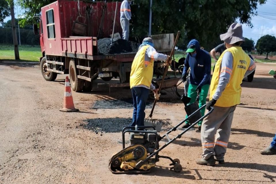 A foto mostra homens trabalhando em serviços de tapa-buracos, aplicando o material na via e um caminhão ao fundo, apoiando a obra