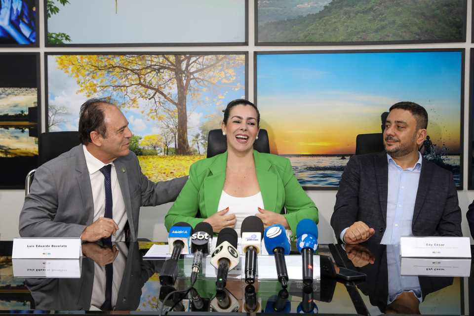 Mulher aparece sentada em uma cadeira, acompanhada de dois homens sentados ao seu lado