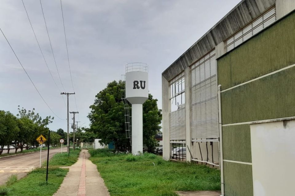 A imagem mostra um reservatório de água alto, pintado de branco, com as letras “RU” em preto na parte frontal. Ele possui uma escada metálica externa que leva até a parte superior, onde há um pequeno guarda-corpo circular. O reservatório fica ao lado de um prédio grande de fachada verde e cinza, com janelas amplas. Em primeiro plano, há uma calçada de concreto com uma faixa tátil vermelha ao centro, cercada por grama alta. À esquerda, vê-se uma rua com postes de energia, árvores e algumas placas de trânsito. O céu está nublado, dando um tom cinza à cena.
