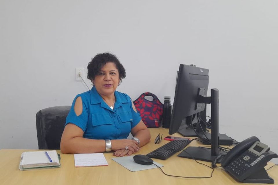 Elisana Garcia, mulher de cabelos curtos e cacheados, vestindo blusa azul, está sentada em uma mesa de escritório, em frente a um computador. Sobre a mesa há agenda, caderno, telefone fixo, teclado, mouse e bolsa ao fundo. Ela olha para a câmera e sorri levemente.