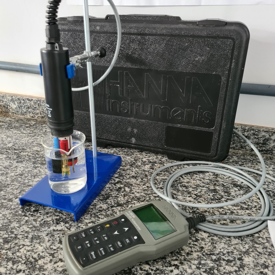 Um equipamento de medição de qualidade da água está sobre uma bancada. Um sensor conectado ao aparelho é mergulhado em um recipiente contendo água, com uma caixa preta da marca "Hanna Instruments" ao fundo.