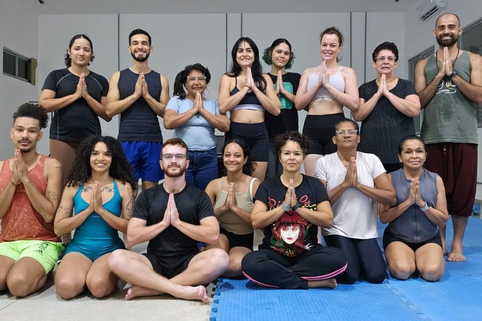Foto de um grupo de participantes reunidos em uma sala, em duas fileiras, com as mãos unidas em frente ao peito, durante uma prática de Yoga.