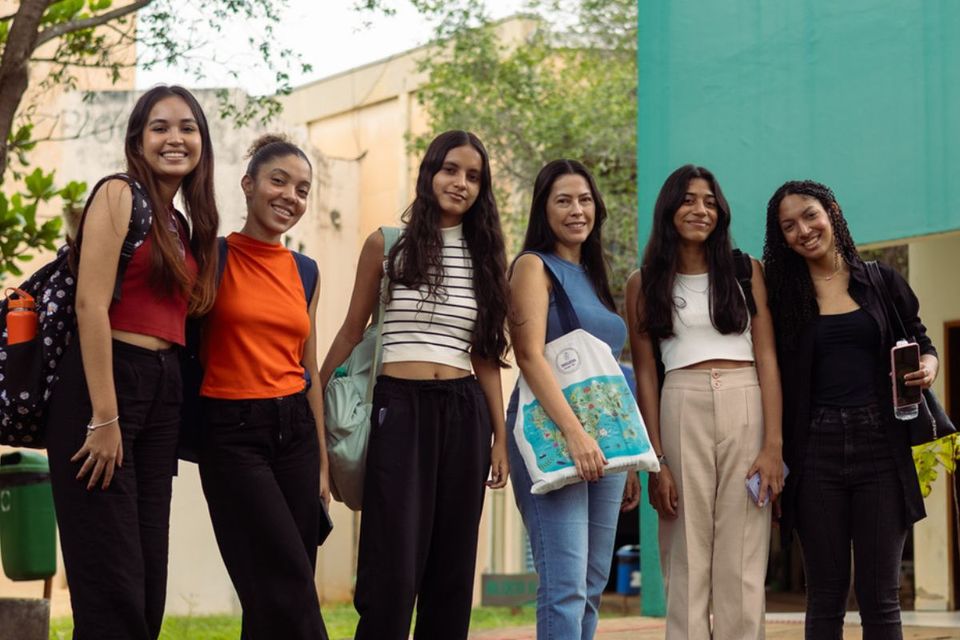 A imagem mostra seis mulheres em pé, sorrindo para a câmera, em um espaço aberto do Câmpus de Palmas da UFT. Elas carregam mochilas e bolsas, aparentando ser estudantes. Ao fundo, há árvores, um prédio de cor clara e uma parede verde. A cena transmite leveza e acolhimento no ambiente universitário.
