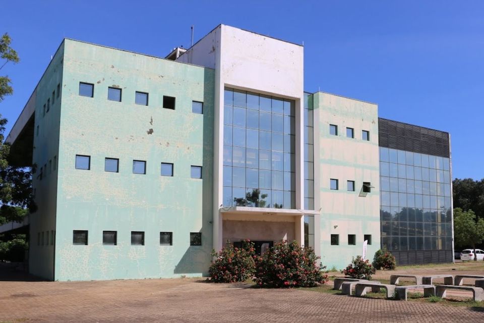 Imagem colorida da fachada da Biblioteca Prof. José Torquato Carolino, no Câmpus Palmas da UFT. O prédio é de três andares, com estrutura retangular, paredes em tom verde-claro desgastado e uma grande área central com vidraças espelhadas. Há canteiros com arbustos floridos em frente à entrada principal e bancos de concreto dispostos na área externa pavimentada. O céu está azul e sem nuvens.