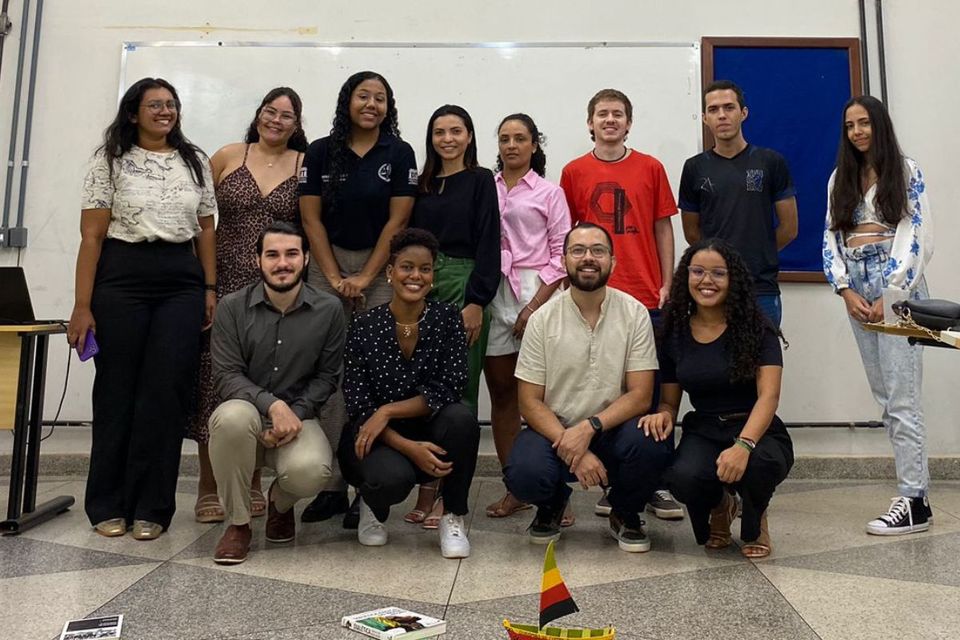 Foto de um grupo de pessoas, algumas em pé, ao fundo, e outras agachadas, à frente, dentro de uma sala de aula, com objetos e livros espalhados pelo chão