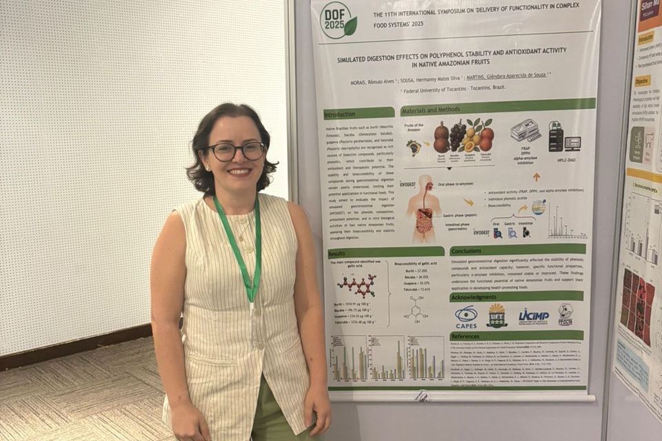 Mulher de cabelos curtos e óculos, usando blusa clara sem mangas e calça verde, sorri diante de um pôster científico afixado em painel branco. O pôster exibe gráficos, diagramas e textos sobre compostos fenólicos e frutos amazônicos, com logotipos da CAPES, UFT e LICIMP na parte inferior.