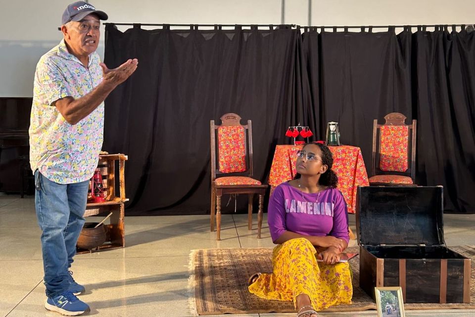 Idoso em pé de boné olhando em direção a plateia, enquanto uma jovem permanece ao seu lado sentada no chão