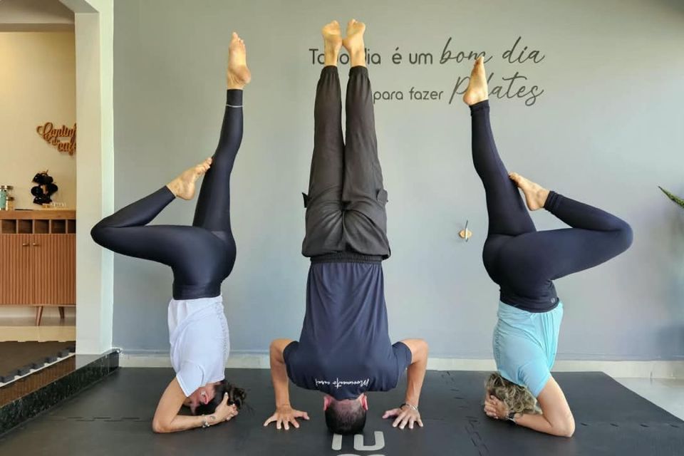 Três pessoas realizam uma postura invertida de Yoga sobre colchonetes, com as pernas elevadas e apoiadas na posição vertical.
