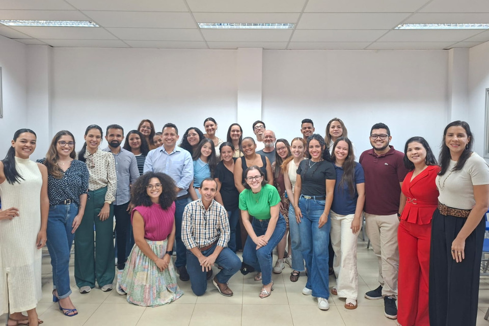 Fotografia colorida de um grupo de aproximadamente 25 pessoas reunidas em uma sala, posando para a foto. Os participantes estão organizados em três fileiras e sorriem para a câmera.