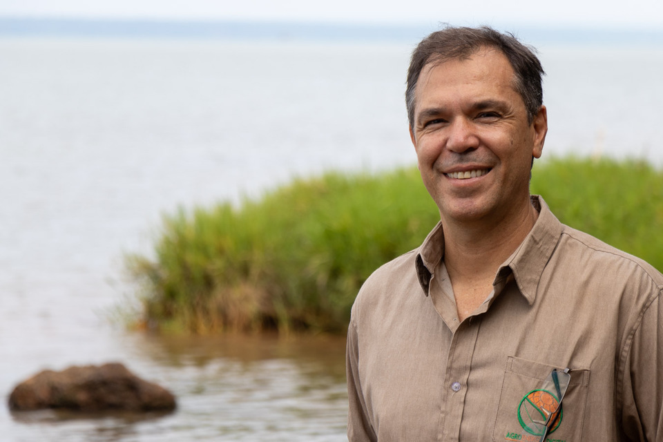 Homem de cabelo grisalho sorri para foto com lago ao fundo
