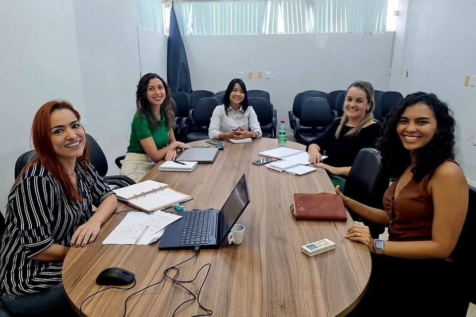 Foto de um grupo de mulheres sentadas em uma mesa de reuniões, olhando e sorrindo para a câmera