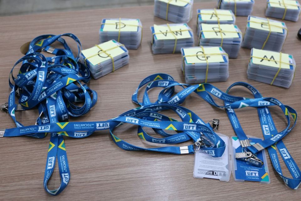 Sobre uma mesa de madeira, há diversos crachás empilhados e amarrados com elásticos, identificados por letras em papéis amarelos. À frente, estão espalhados vários cordões azuis com o logotipo e o nome da Universidade Federal do Tocantins impressos em branco, junto a alguns crachás prontos para uso.