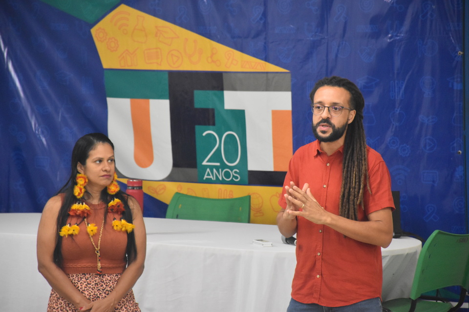 Imagem colorida com duas pessoas e uma mesa e um fundo com a logo da UFT