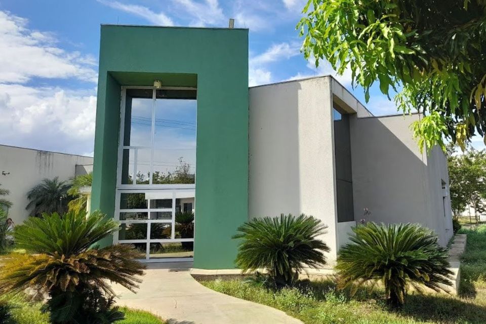 Fotografia da fachada do prédio da Direção do Câmpus de Palmas da UFT. O edifício possui paredes nas cores verde e branca, grandes janelas de vidro e uma entrada cercada por plantas e árvores. Ao fundo, o céu está azul, com algumas nuvens.