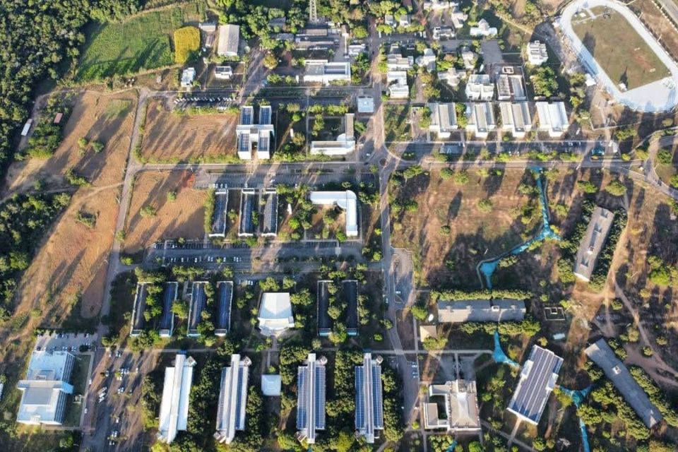 A imagem mostra uma vista aérea do câmpus de Palmas, com vários prédios brancos e de telhados azulados, dispostos em fileiras entre ruas internas e áreas verdes. Há árvores espalhadas por todo o local e algumas áreas de terreno mais seco. No canto superior direito, vê-se uma pista ovalada com gramado ao centro, provavelmente uma pista de atletismo ou campo esportivo. As sombras dos prédios e das árvores são longas, indicando que a foto foi tirada no início da manhã ou no fim da tarde. O câmpus está cercado por vegetação e pequenas áreas rurais ao fundo.