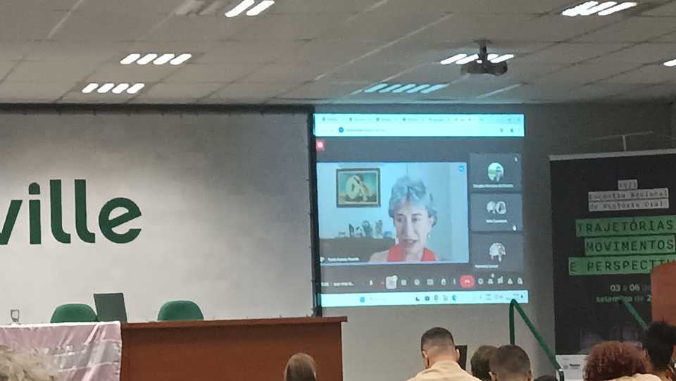 Imagem colorida com foto da professora no telão, participando da entrega online