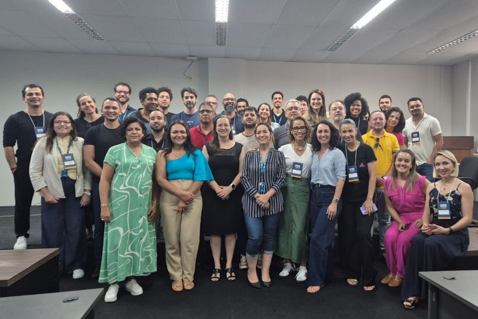 Foto colorida em ambiente interno mostra cerca de 30 pessoas reunidas em grupo, posando para a câmera. Elas estão organizadas em duas fileiras, uma em pé ao fundo e outra à frente. A maioria utiliza crachá de identificação. O espaço é uma sala de reuniões com iluminação branca no teto, paredes claras e mesas nas laterais. As pessoas vestem roupas variadas, desde trajes mais formais até casuais, e demonstram expressão de satisfação, muitas sorrindo.