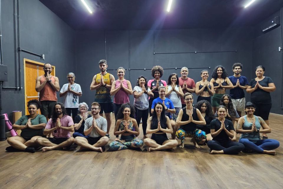 Um grupo diverso de pessoas posa sentado e em pé em sala de prática de Yoga, com as mãos unidas em posição de saudação, formando uma grande turma sorridente.