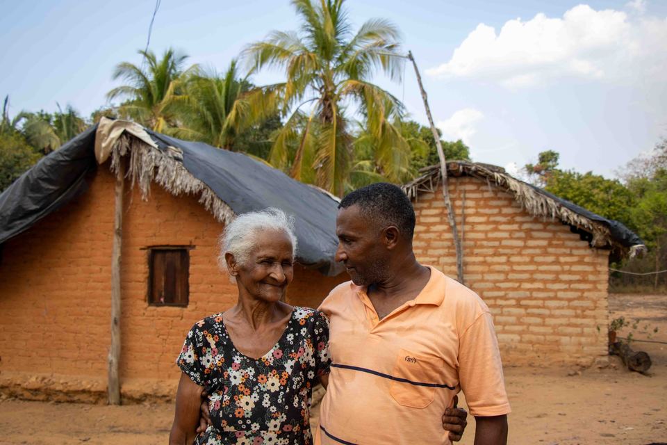 uma senhora e um homem negros diante de uma casa de adobe
