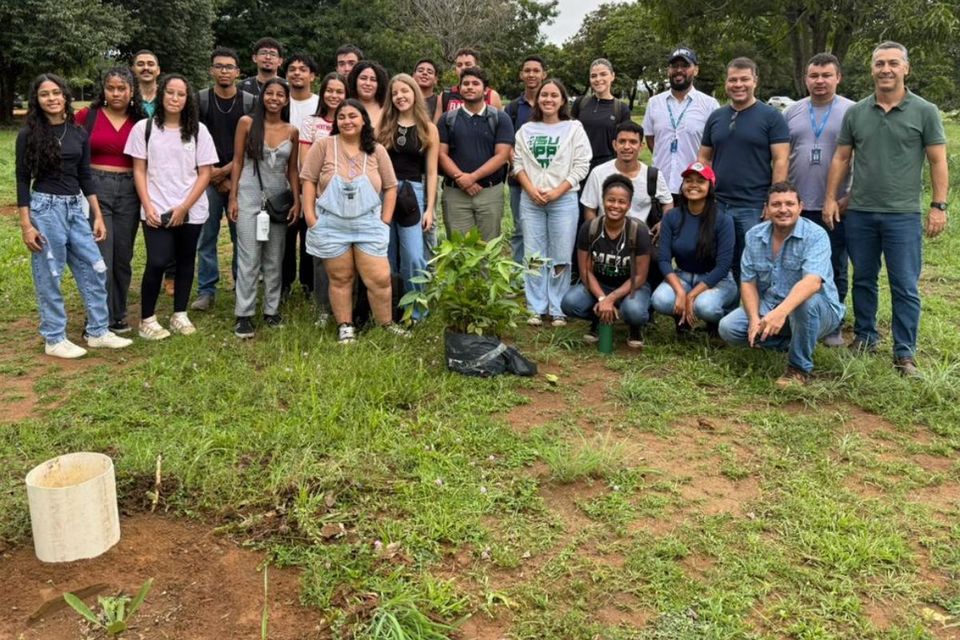 Fotografia colorida mostra um grupo de aproximadamente 25 pessoas reunidas em uma área gramada e arborizada ao ar livre. Entre os participantes há estudantes, professores e servidores, posicionados em pé e agachados para o registro coletivo. No centro da imagem, em primeiro plano, aparecem mudas de árvores preparadas para plantio. Ao fundo, há árvores de grande porte e vegetação, compondo um ambiente natural. 