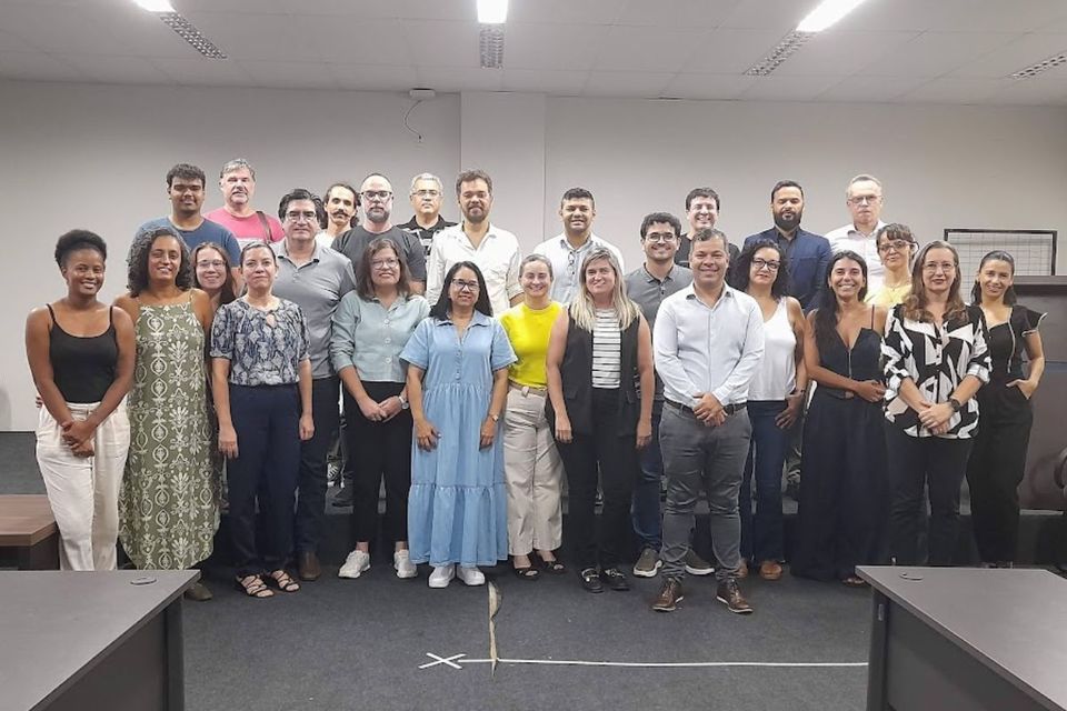 A imagem mostra um grupo de aproximadamente 27 pessoas, entre homens e mulheres, posando para uma foto em um ambiente fechado, possivelmente uma sala de reuniões ou auditório. Todos estão em pé, organizados em três fileiras, e olham para a câmera com expressões amigáveis. A iluminação é clara, e o ambiente tem aparência profissional, com teto de forro, luminárias fluorescentes e mesas ao redor. As pessoas estão vestidas com roupas casuais ou sociais leves.