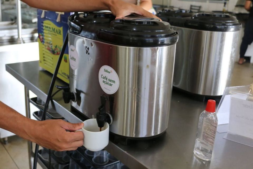 Pessoa serve café em uma xícara branca, utilizando uma torneira de um recipiente térmico de inox. Ao fundo, há outros recipientes semelhantes, identificados para diferenciar as bebidas disponíveis sobre um balcão.