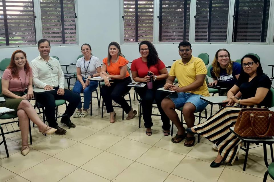 Na foto, oito pessoas sentadas em cadeiras de sala de aula, dispostas em semicírculo. Ao fundo, janelas com persianas. Iluminação artificial no teto.