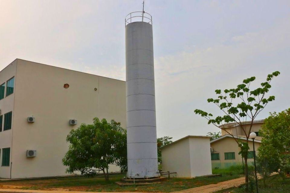 Imagem externa de uma torre cilíndrica branca de armazenamento de água localizada no Câmpus de Palmas da UFT. Ao lado da estrutura há edifícios de cor clara, árvores e áreas gramadas. O céu está parcialmente nublado, e a torre se destaca pela altura em relação às construções ao redor.